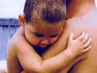 A man at the beach, standing lovingly holding a child in his arms, the child's arm is holding onto back of the man's arm