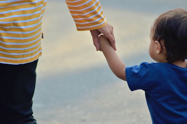 A partial view, seen from the back, of a mother reaching down with her hand to her toddler, as they walk